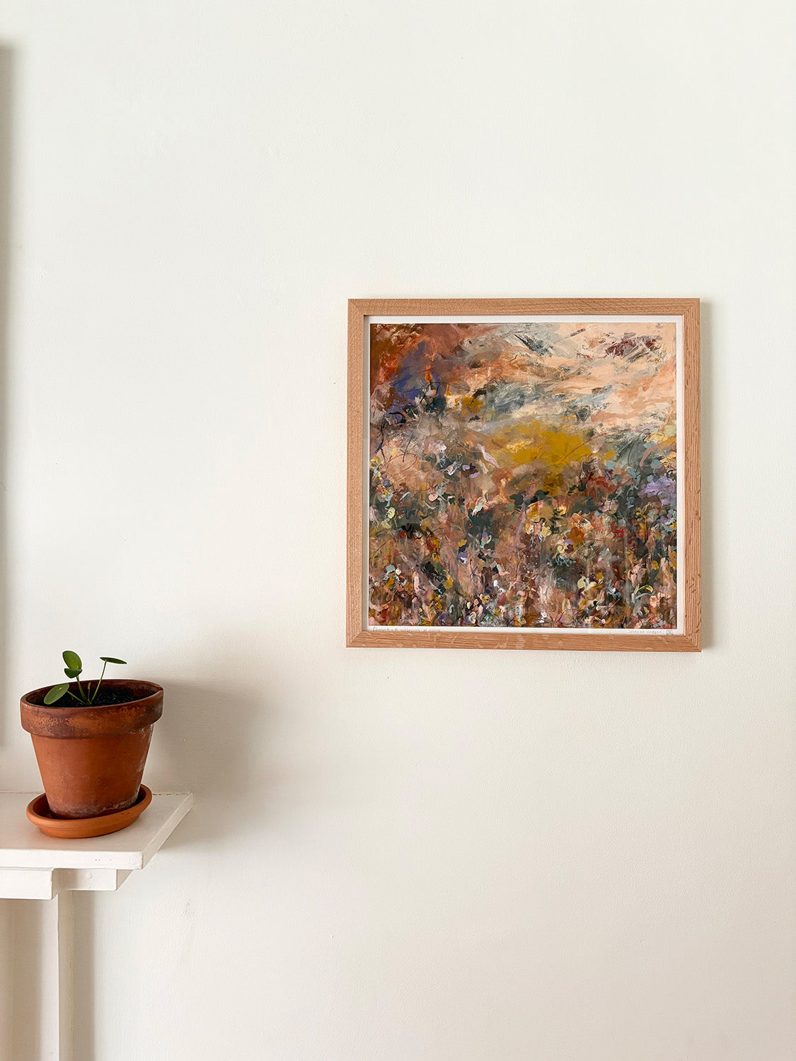 Abstract art print in a wooden frame on a white wall above a mantelpiece with a plant.