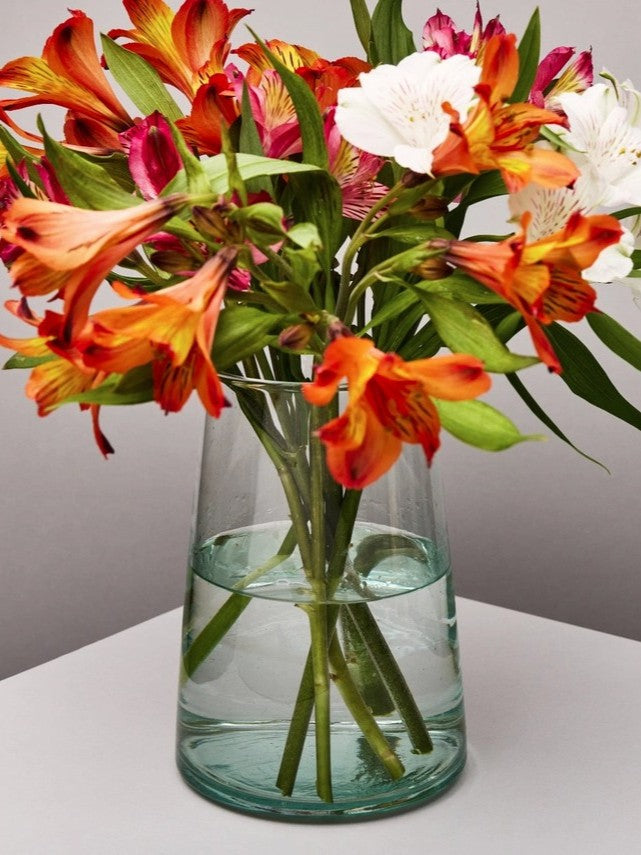Handmade recycled glass tapered vase with colourful flowers on a white surface and neutral background.