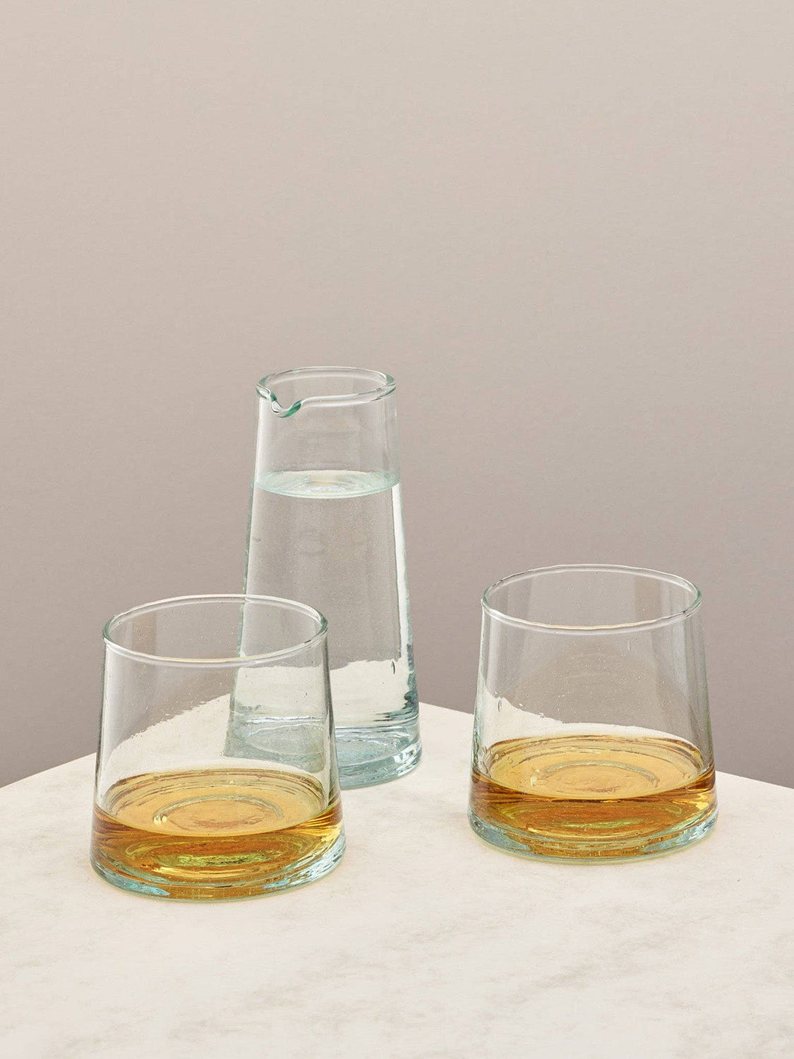 Two glasses with whisky and a small jug on a light surface against a beige background.