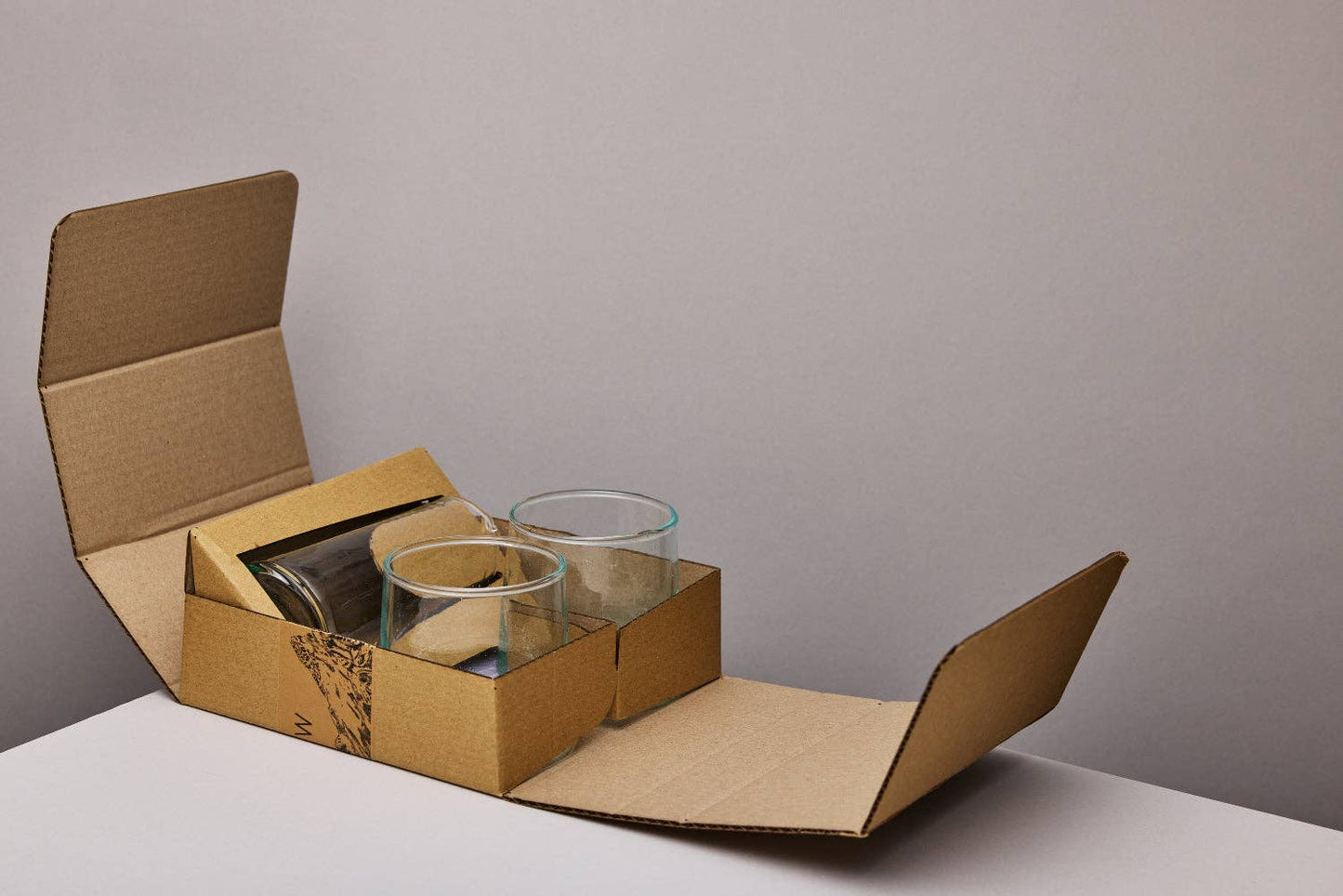 Open cardboard box with glass tumblers and small jug on a neutral background.