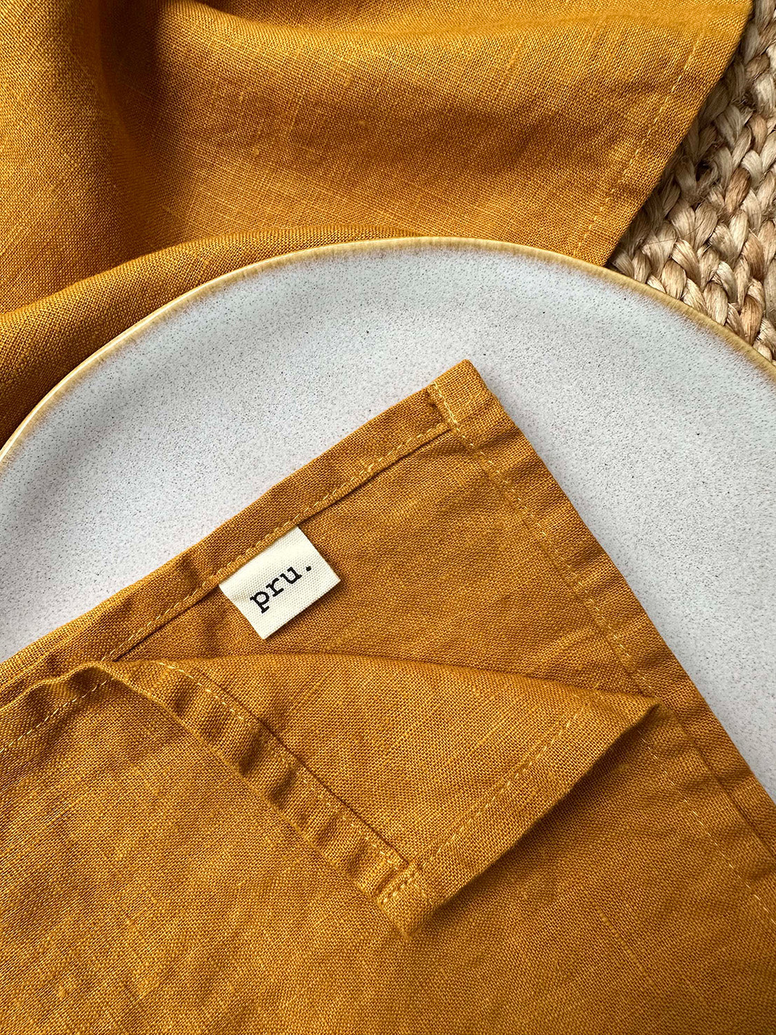 100% linen napkin set in spice colour with 'pru' label, on a white plate with woven placemat.