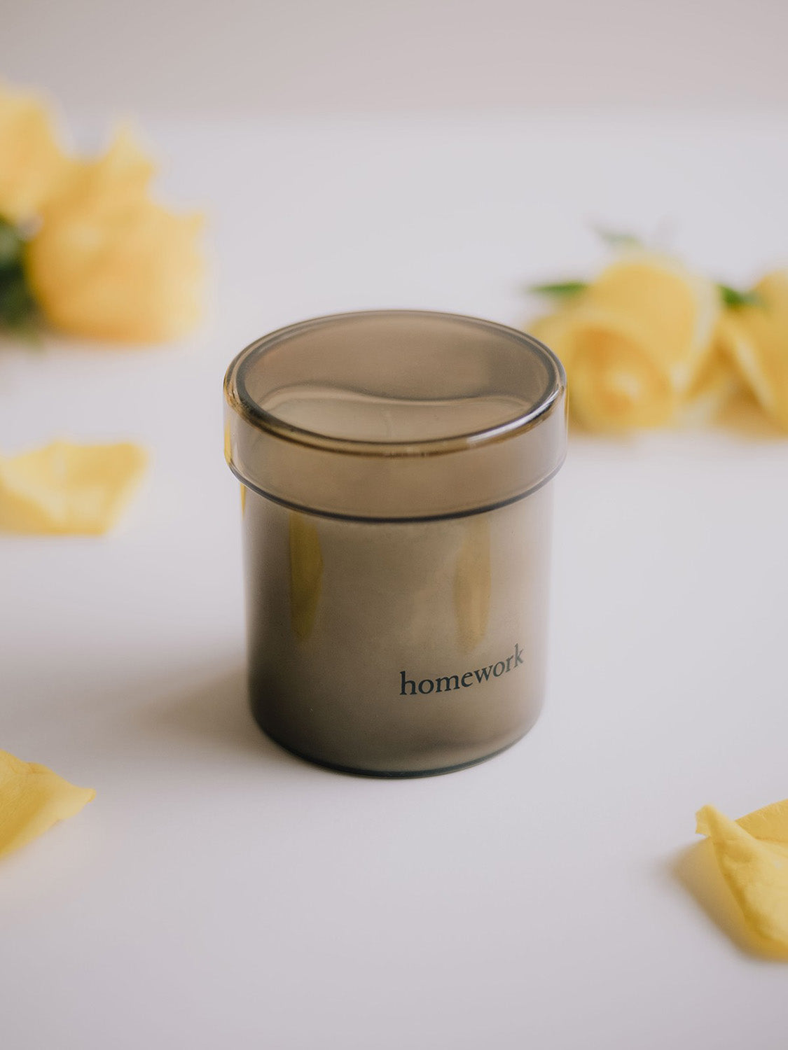 Candle in smoked glass jar labelled 'homework' on a light surface with yellow flowers.