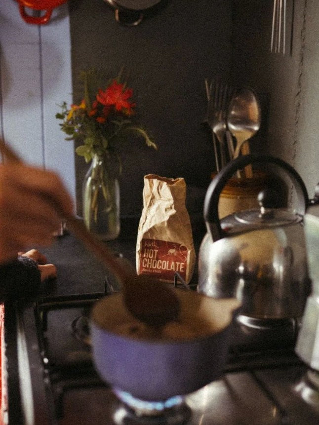 Person stirring a cup of hot chocolate with a spoon on a stove, with a bag of hot chocolate mix and a teapot in the background.