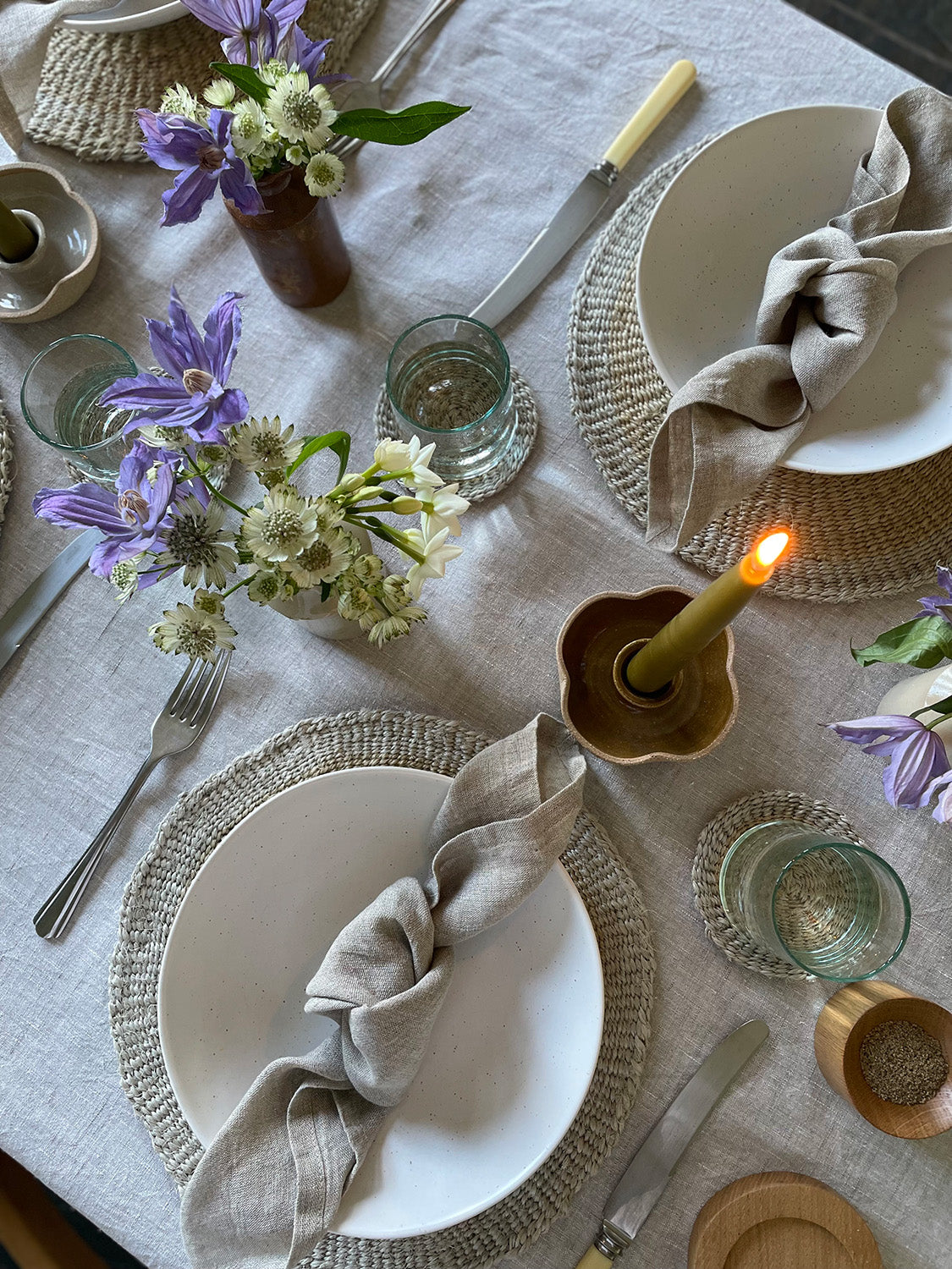 Elegant table setting with white bowls, linen napkins, flowers and candle on a light-coloured tablecloth.