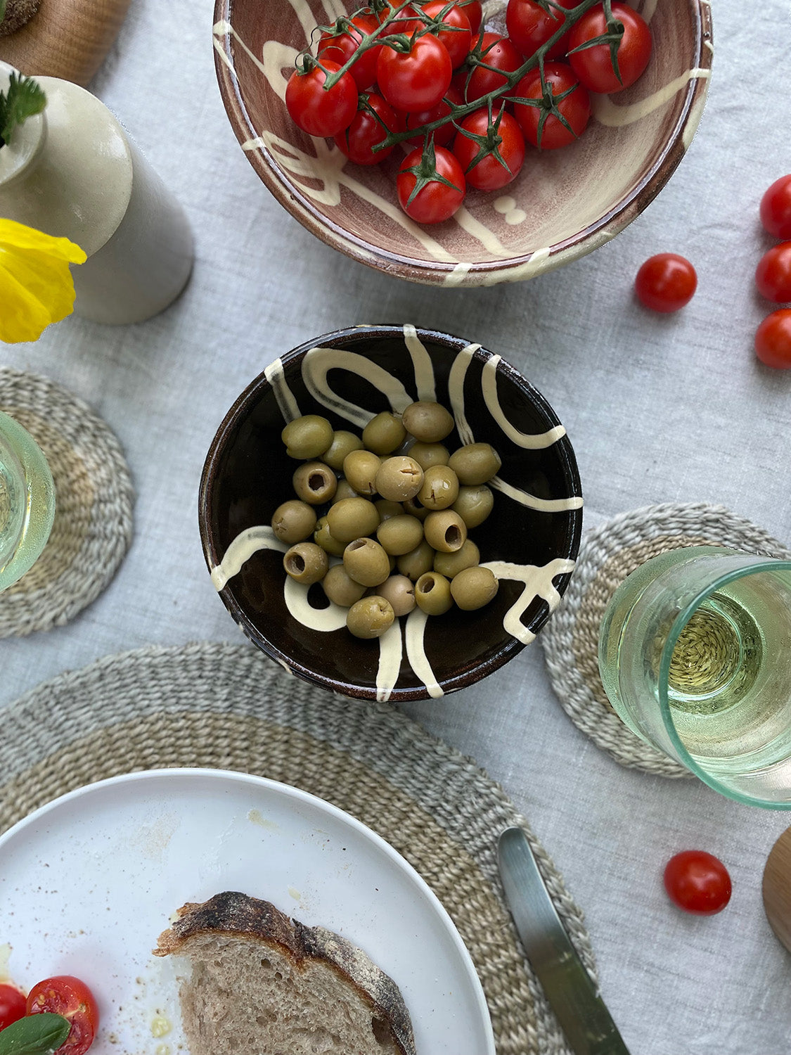 Table setting with a bowl of olives, bread, and tomatoes on a linen tablecloth.