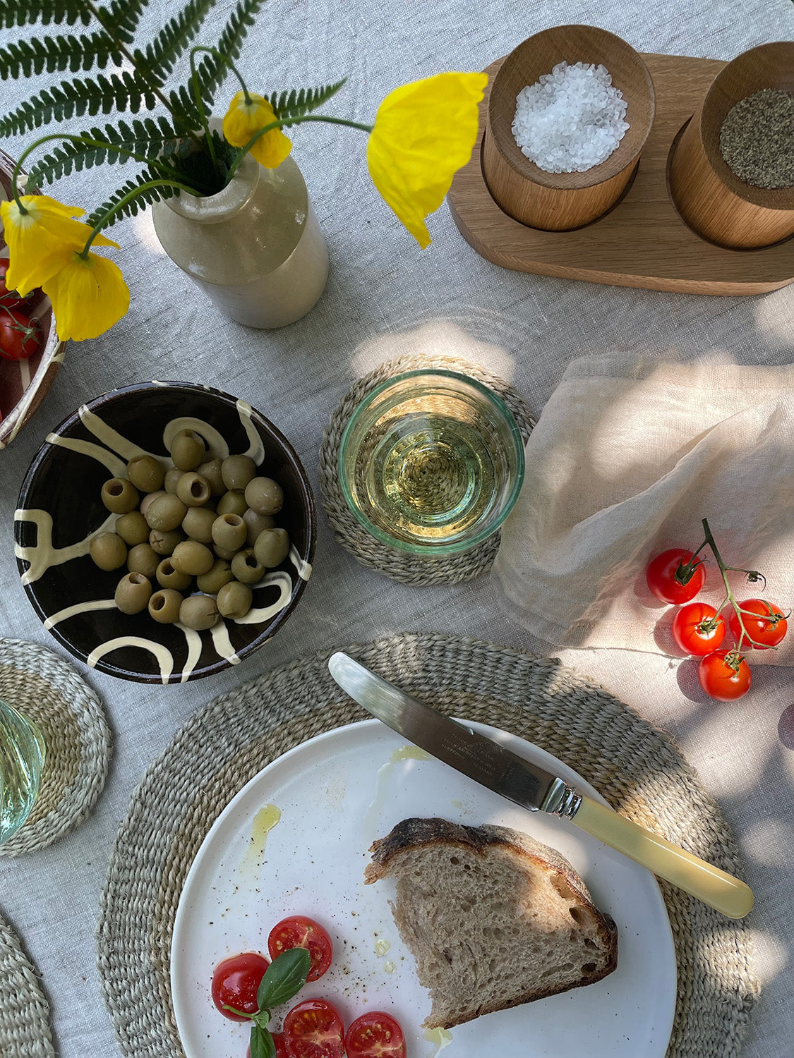 Table setting with bread, tomatoes, olives, and a glass of white wine on a linen tablecloth.