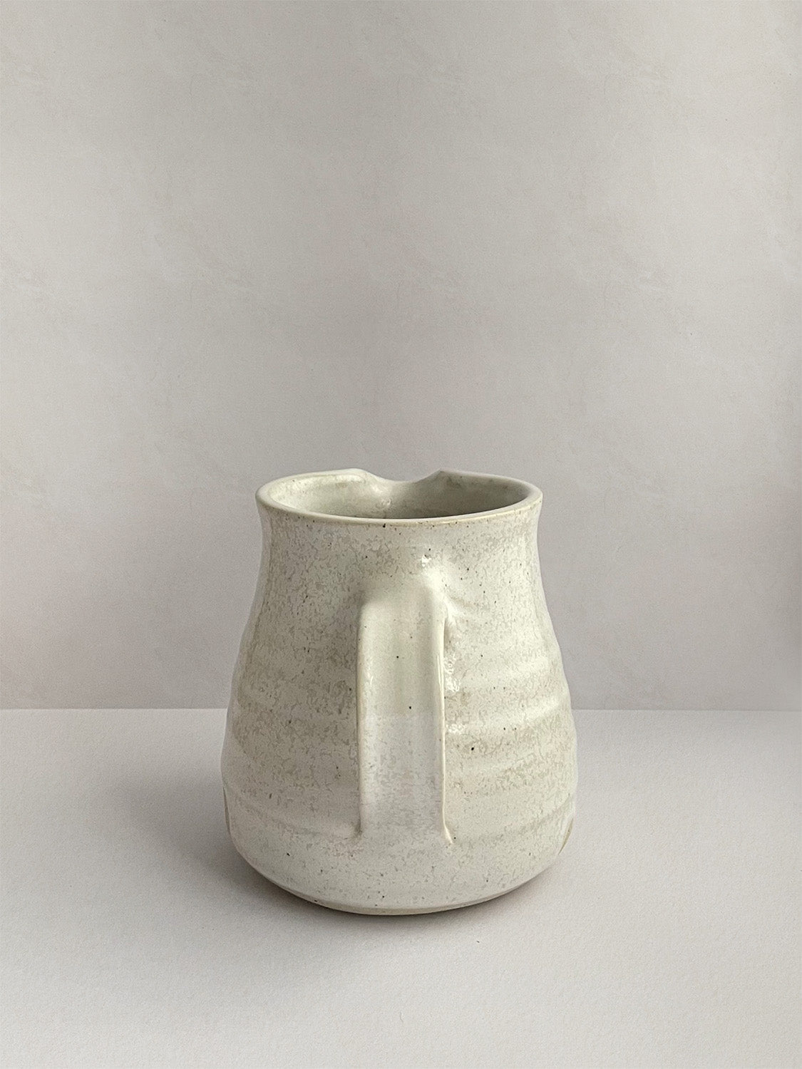A beige handmade ceramic jug with a curved body and a handle, on a neutral background - back view.