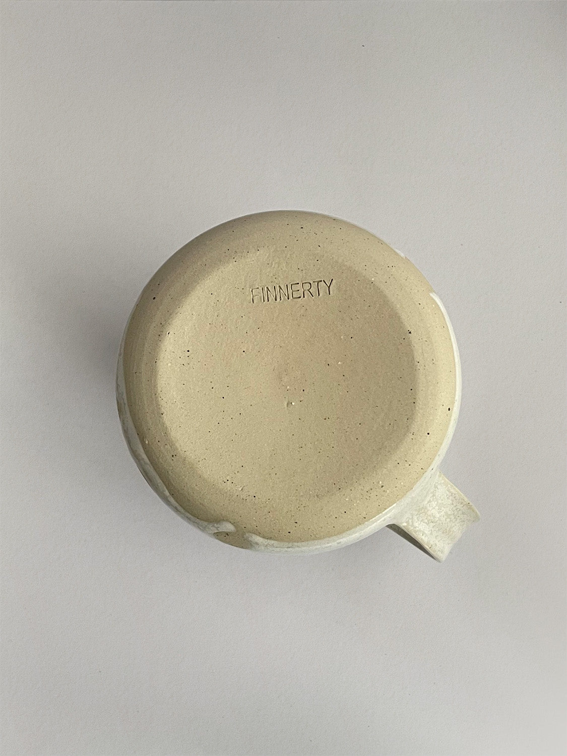 A beige handmade ceramic jug with a curved body and a handle, on a neutral background - base view.