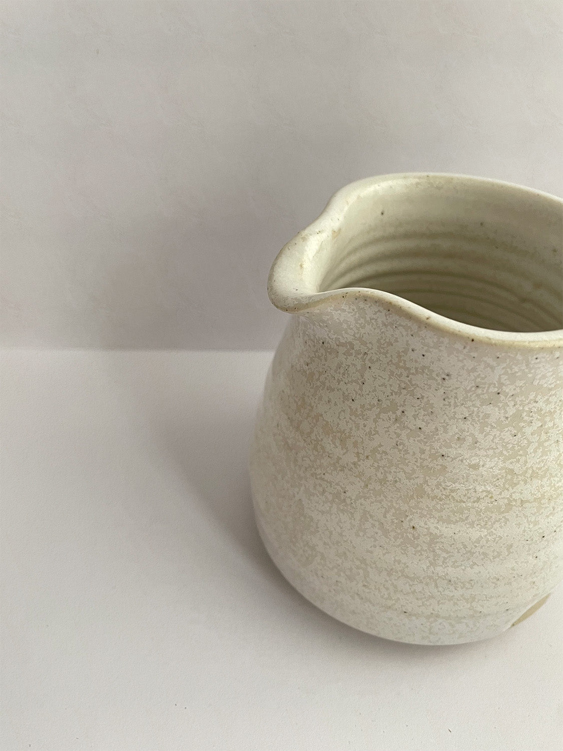 A beige handmade ceramic jug with a curved body and a handle, on a neutral background - detail view.