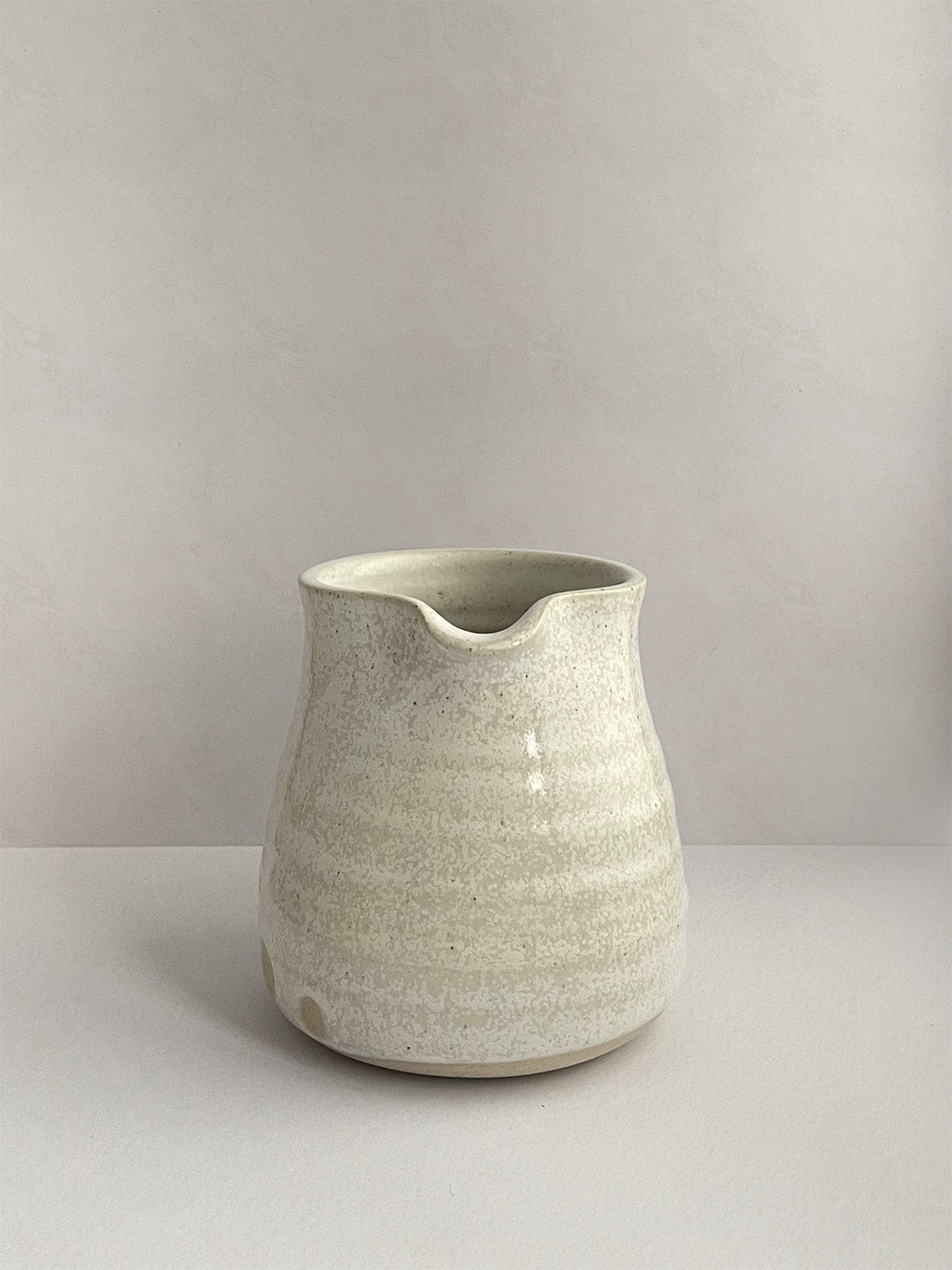 A beige handmade ceramic jug with a curved body and a handle, on a neutral background - front view.