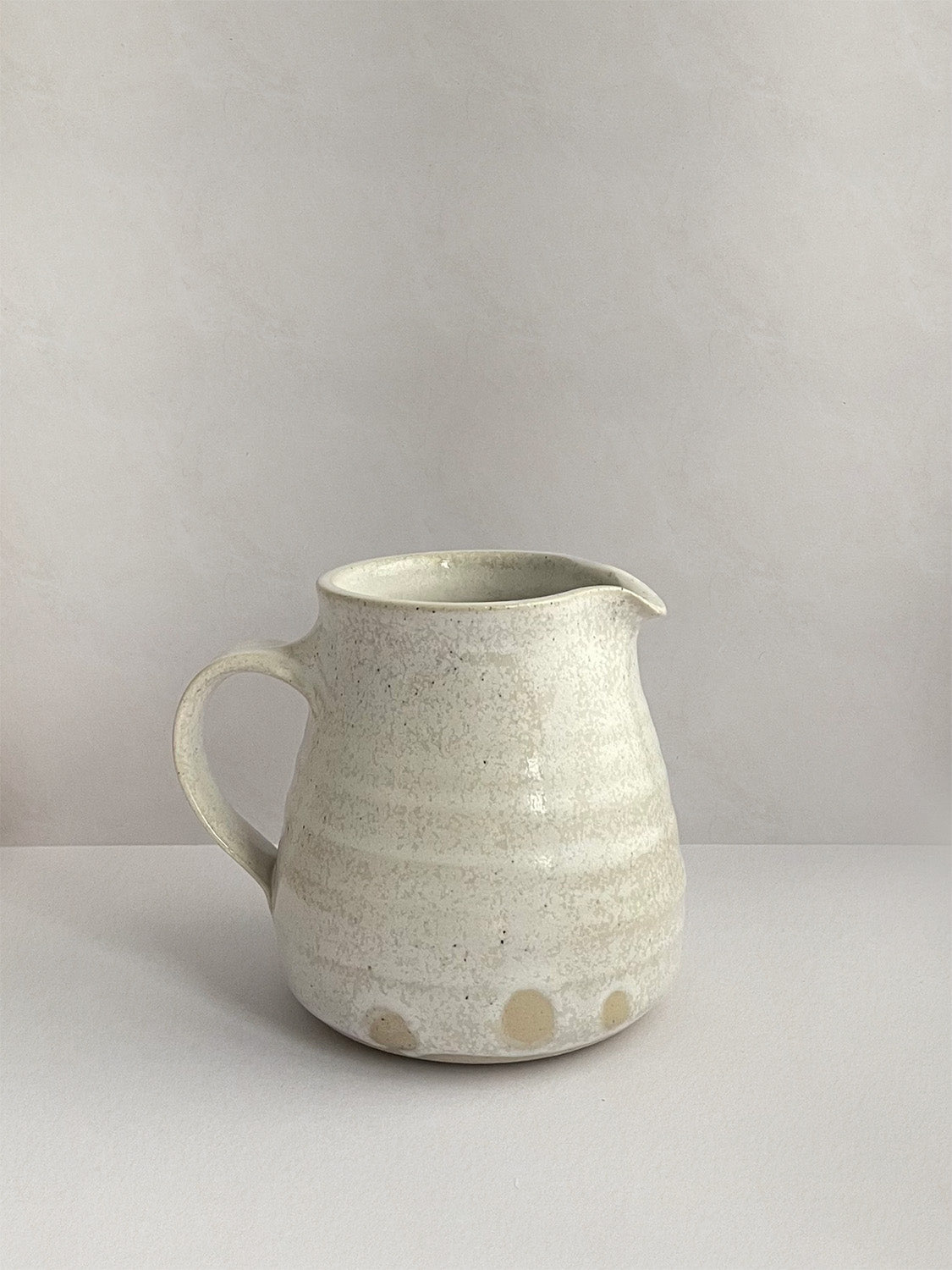 A beige handmade ceramic jug with a curved body and a handle, on a neutral background - right side view.