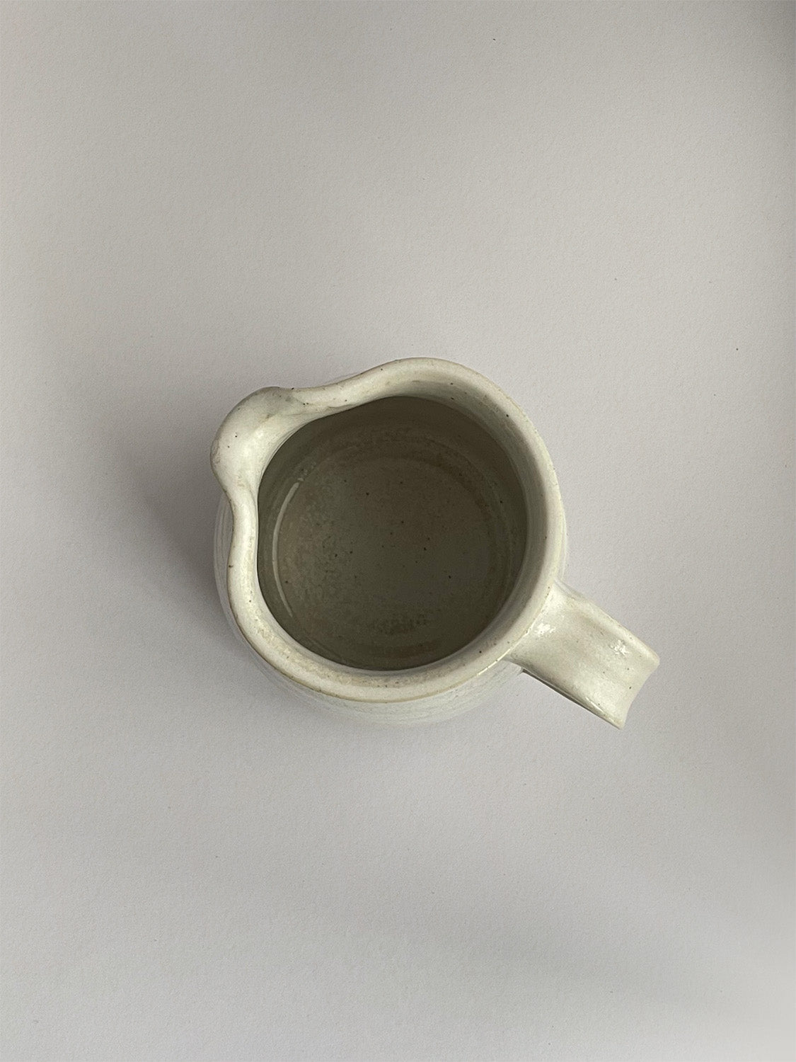 A beige handmade ceramic jug with a curved body and a handle, on a neutral background - top view.