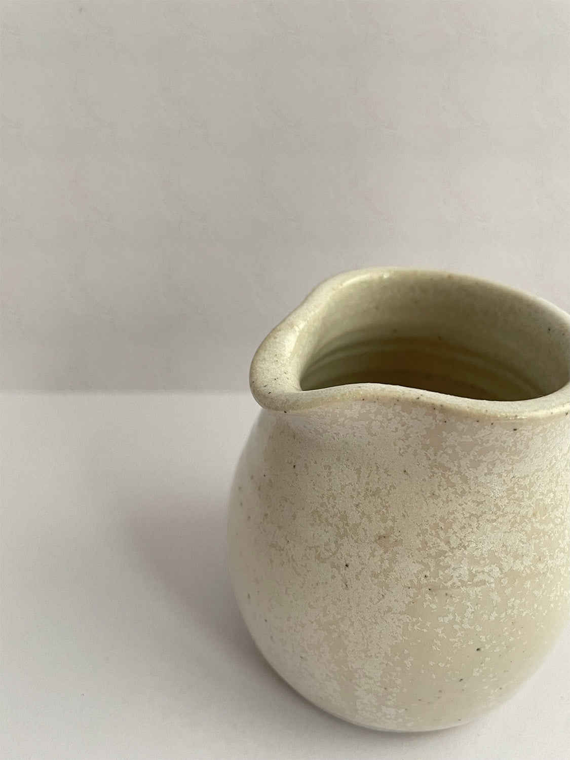 A hand-thrown ceramic milk jug in a beige speckled glaze on a neutral background - close-up view.