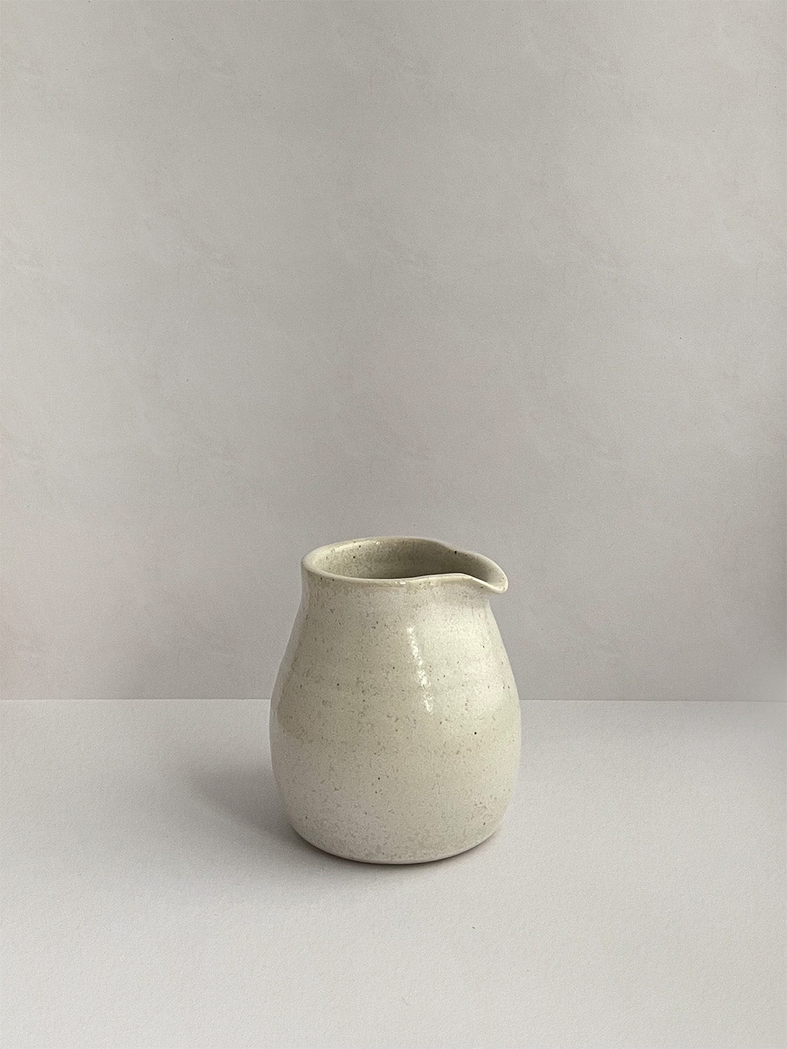 A hand-thrown ceramic milk jug in a beige speckled glaze on a neutral background - right side view.
