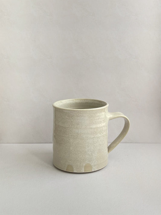 A hand-thrown ceramic mug with a handle, in a beige speckled glaze on a neutral background - left side view.