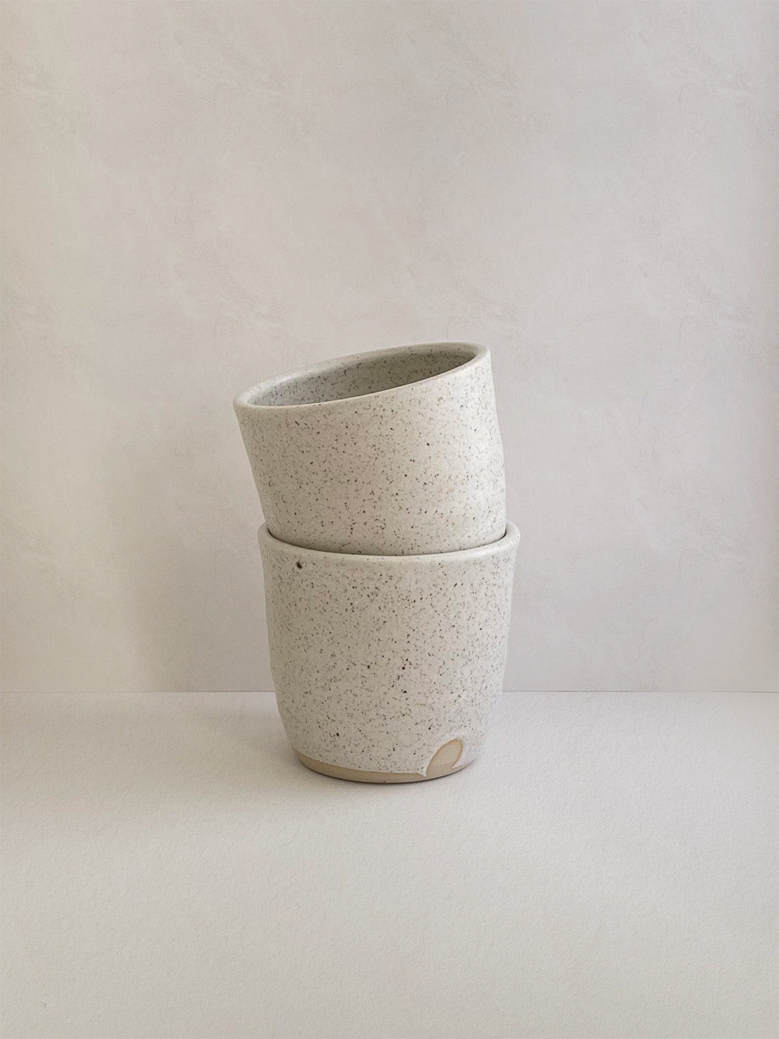 Two stacked white ceramic cups with a speckled texture on a neutral background.