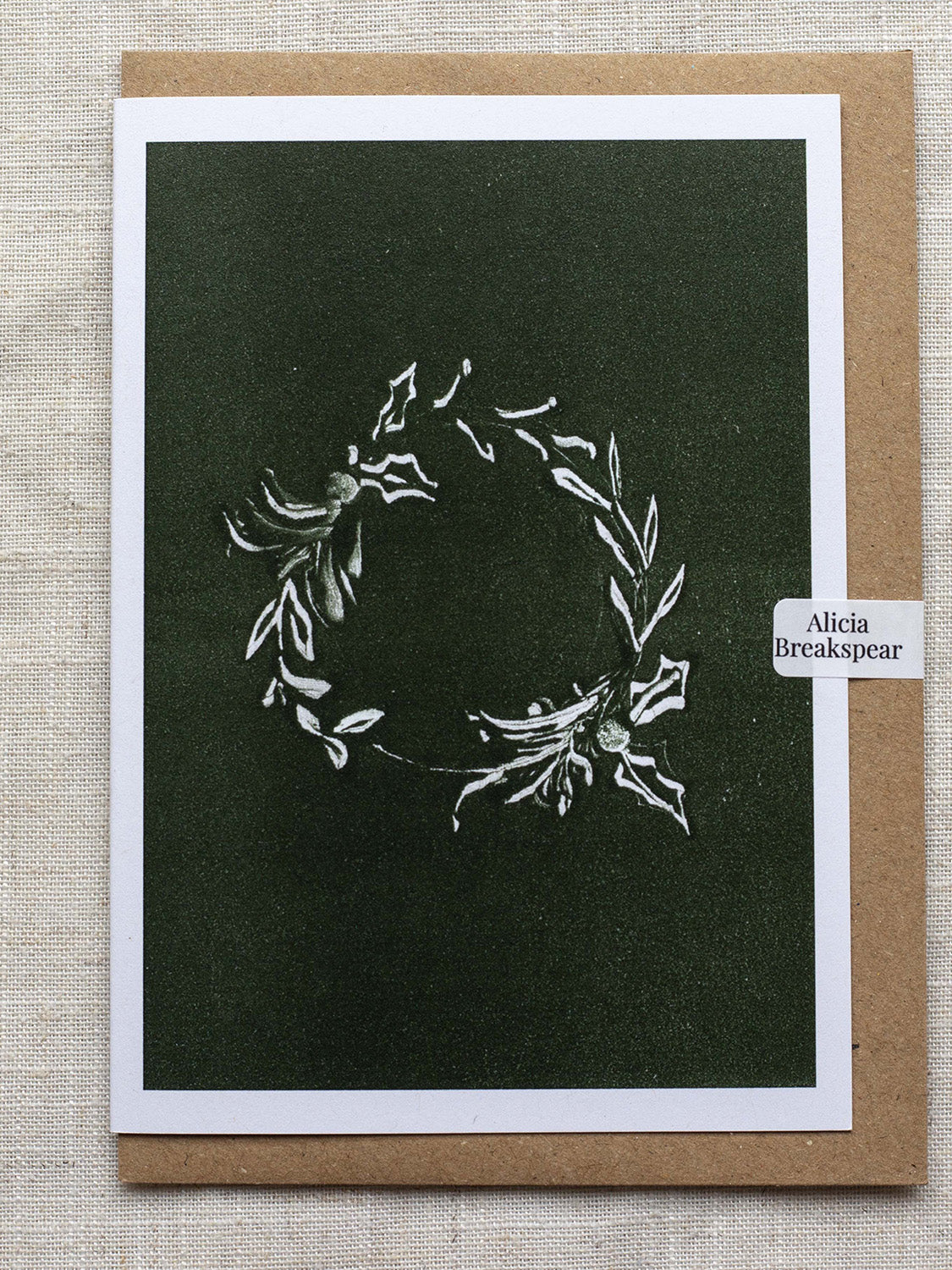 Botanical Christmas card with a leafy wreath design on a green background, with a Kraft paper envelope and 'Alicia Breakspear' sticker seal.