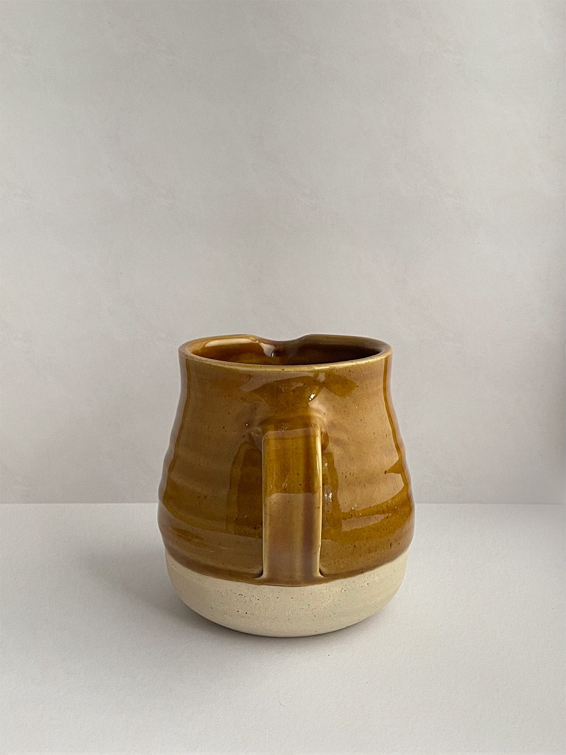 A golden yellow handmade ceramic jug with a curved body and a handle, on a neutral background - back view.