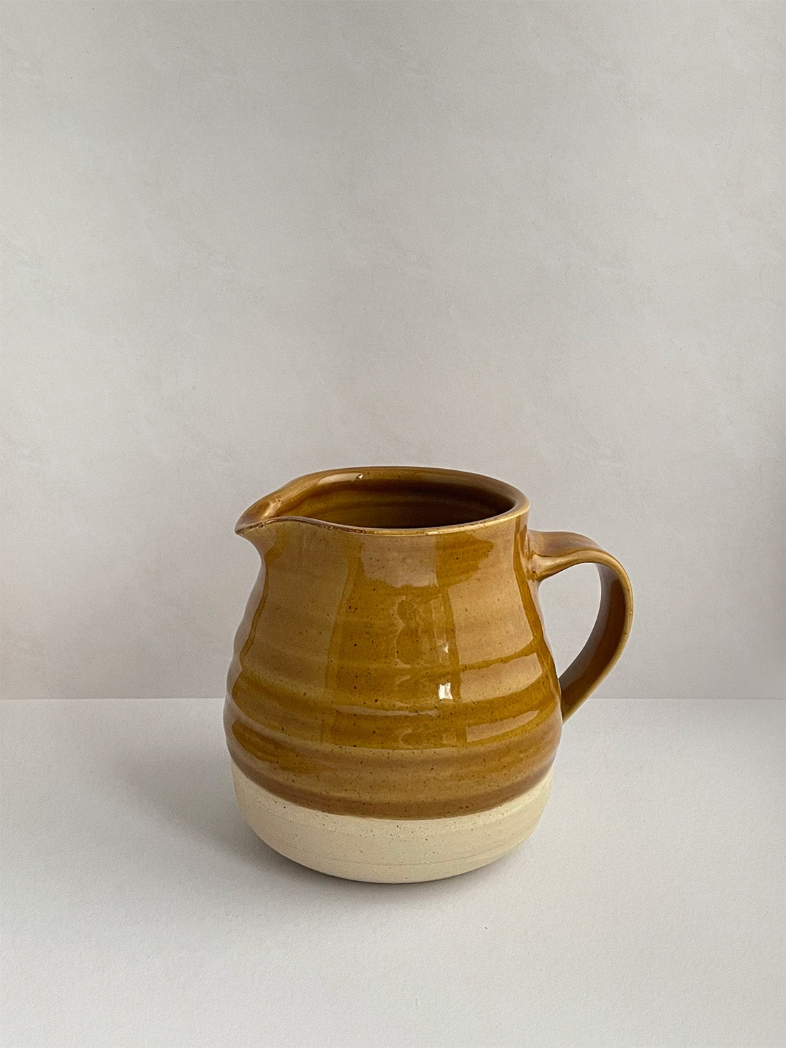 A golden yellow handmade ceramic jug with a curved body and a handle, on a neutral background - left side view.
