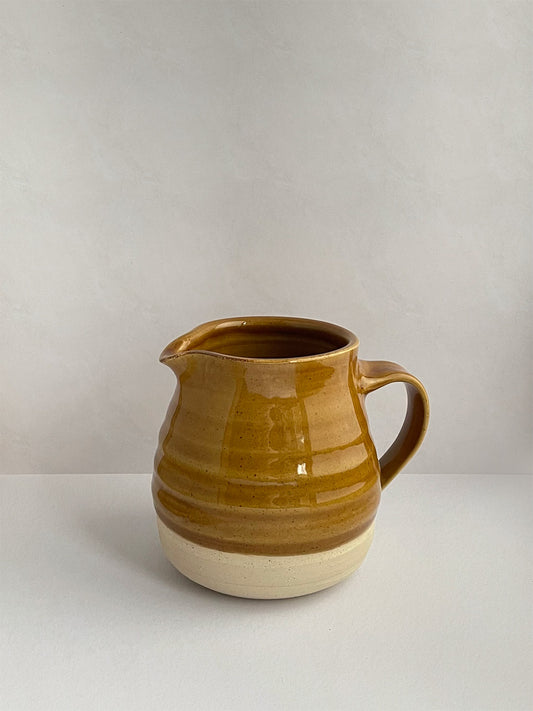 A golden yellow handmade ceramic jug with a curved body and a handle, on a neutral background - left side view.