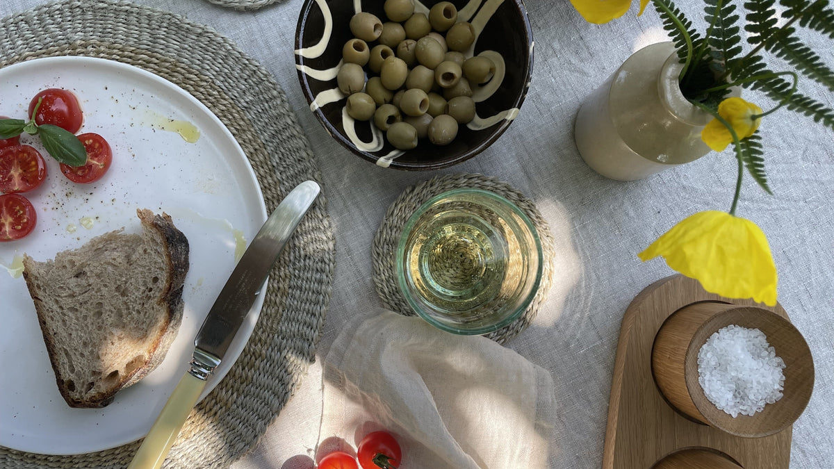 Sunlit summer table setting with olives, tomatoes, bread, wine and handmade tableware in a garden setting.
