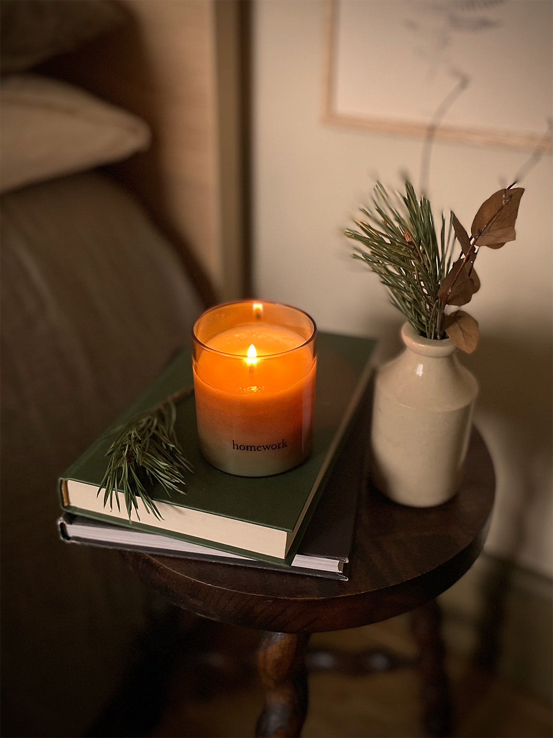 Candle on a stack of books with a vase of greenery on a bedside table in a cosy room.