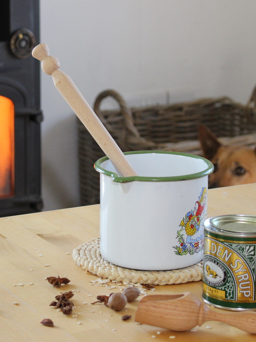 Enamel pot with beech wood porridge spurtle on a table near a can of syrup in a cosy kitchen setting.