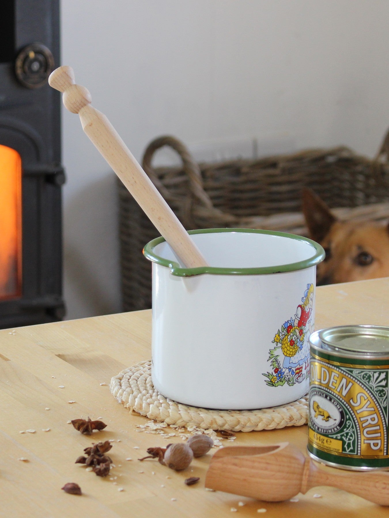 Enamel pot with beech wood porridge spurtle on a table near a can of syrup in a cosy kitchen setting.