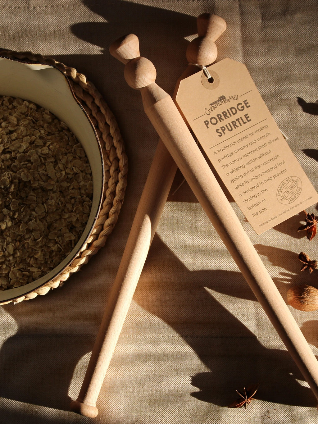 Two beech wood porridge spurtles  with a Kraft paper tag on a textured surface with a pan of oats.