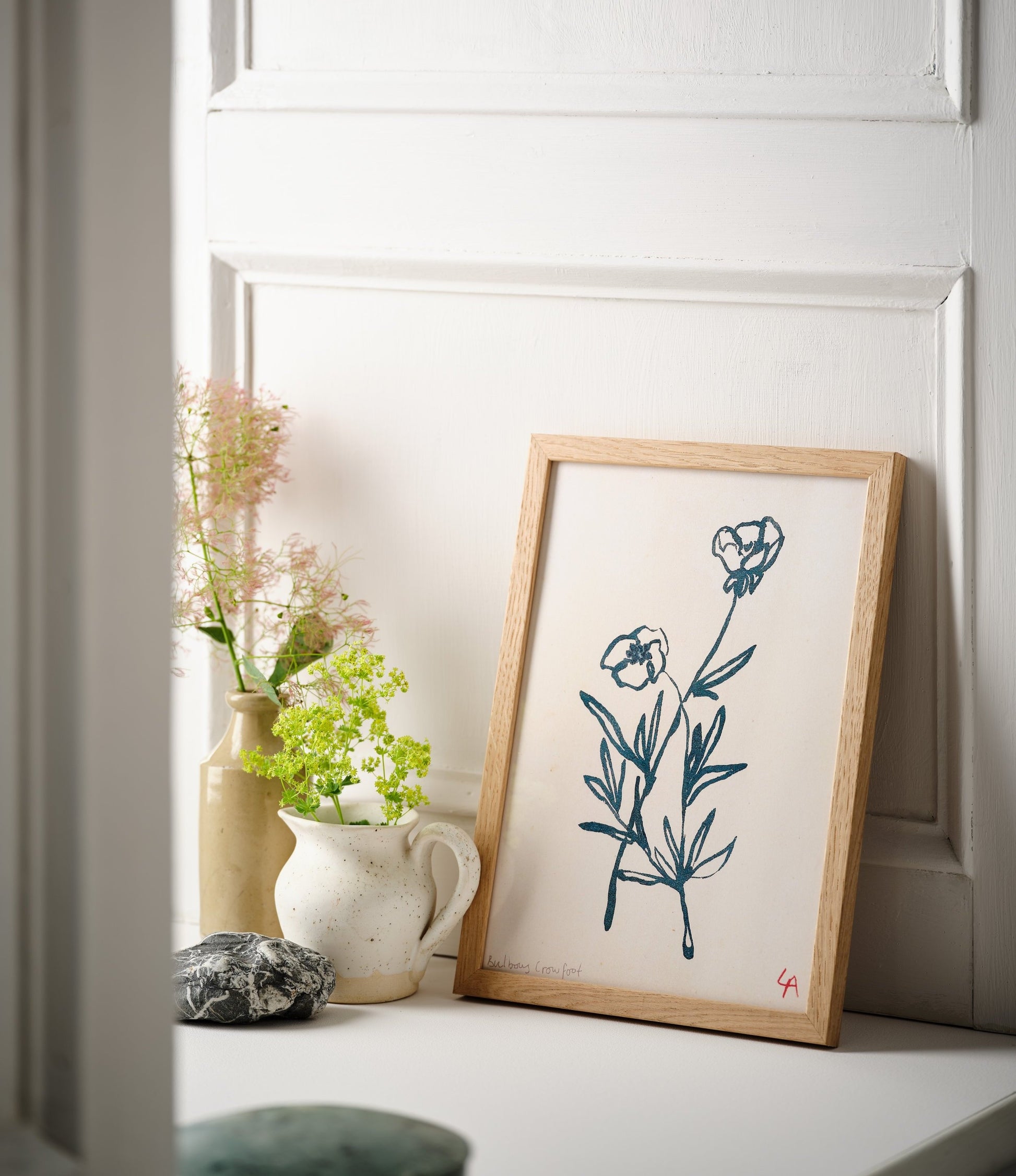 Art print of two Bulbous Crowfoot flowers in teal ink on vintage paper in a natural oak frame, sitting on a white painted windowsill alongside two stoneware vessels with wildflowers and a large dark grey pebble