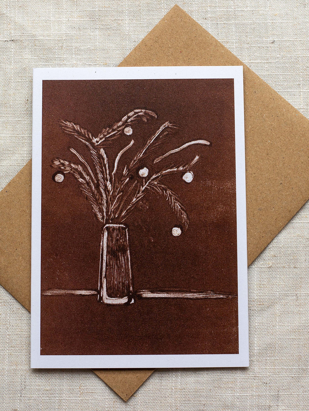 Botanical Christmas card with a vase of festive foliage design on a dark red background, on a Kraft paper envelope placed on a linen surface.