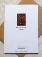 Reverse side of botanical Christmas card with a vase of festive foliage design on a dark red background, on a Kraft paper envelope placed on a linen surface.