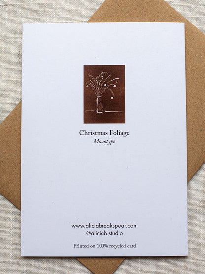 Reverse side of botanical Christmas card with a vase of festive foliage design on a dark red background, on a Kraft paper envelope placed on a linen surface.