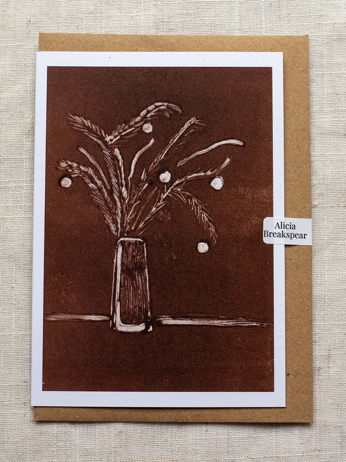 Botanical Christmas card with a vase of festive foliage design on a dark red background, with a Kraft paper envelope and 'Alicia Breakspear' sticker seal.