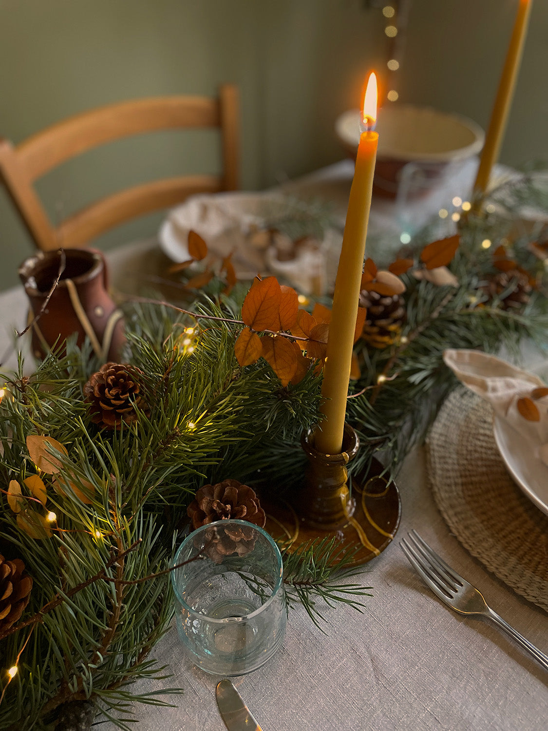 Decorative table setting with candles, greenery, and pinecones on a linen tablecloth.