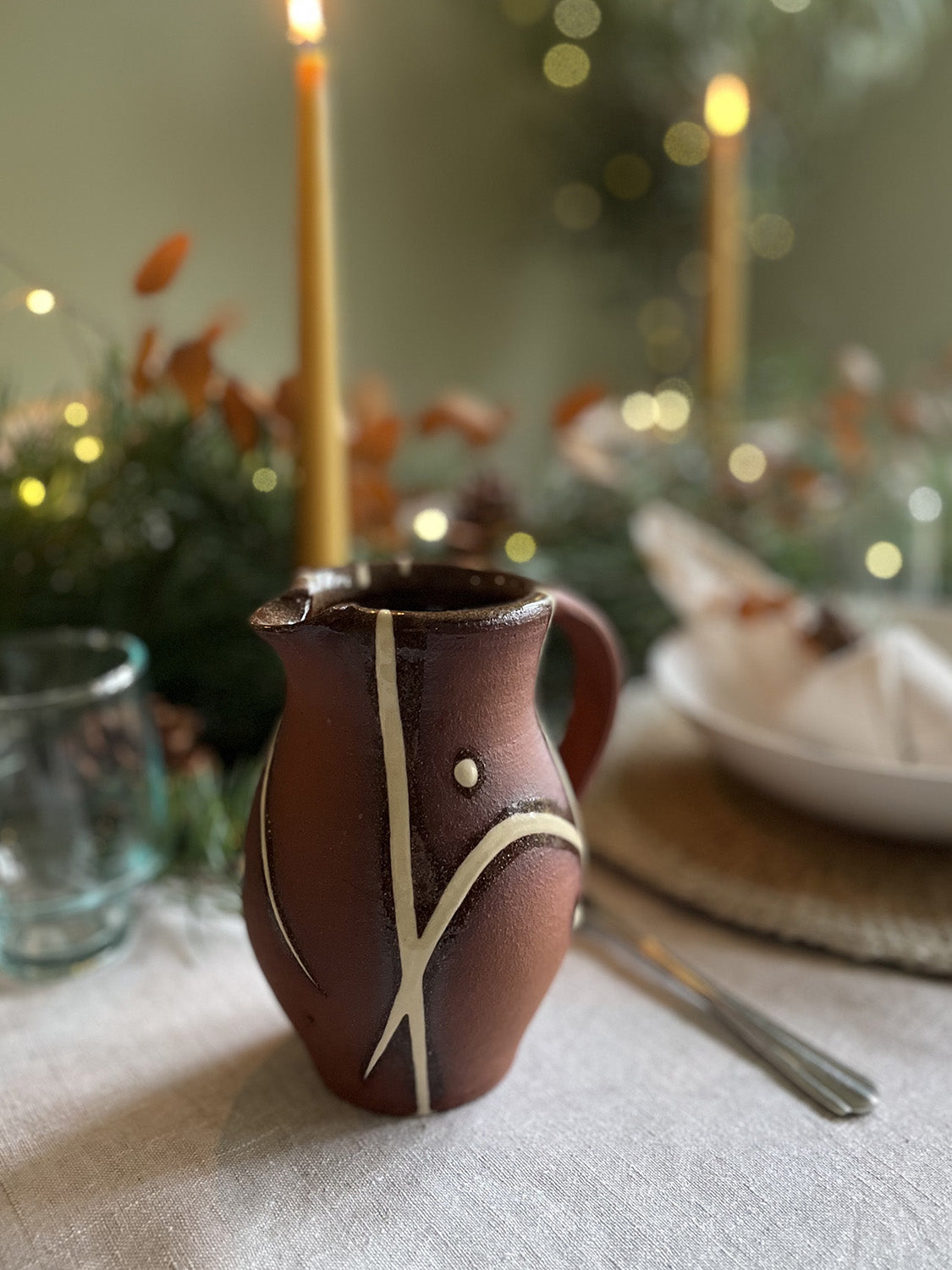 Ceramic pitcher with decorative lines on a table setting with candles and a foliage.