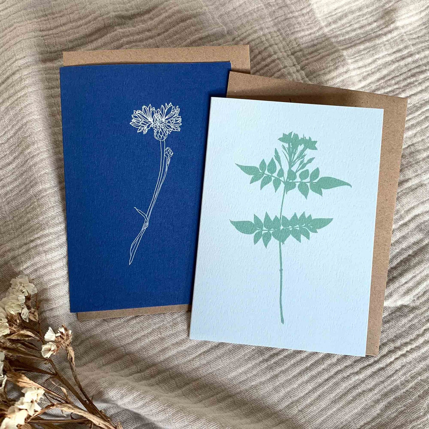 Cornflower and Jasmine botanical greetings cards with Kraft paper envelopes on a cream, rippled fabric background with some dried flowers.