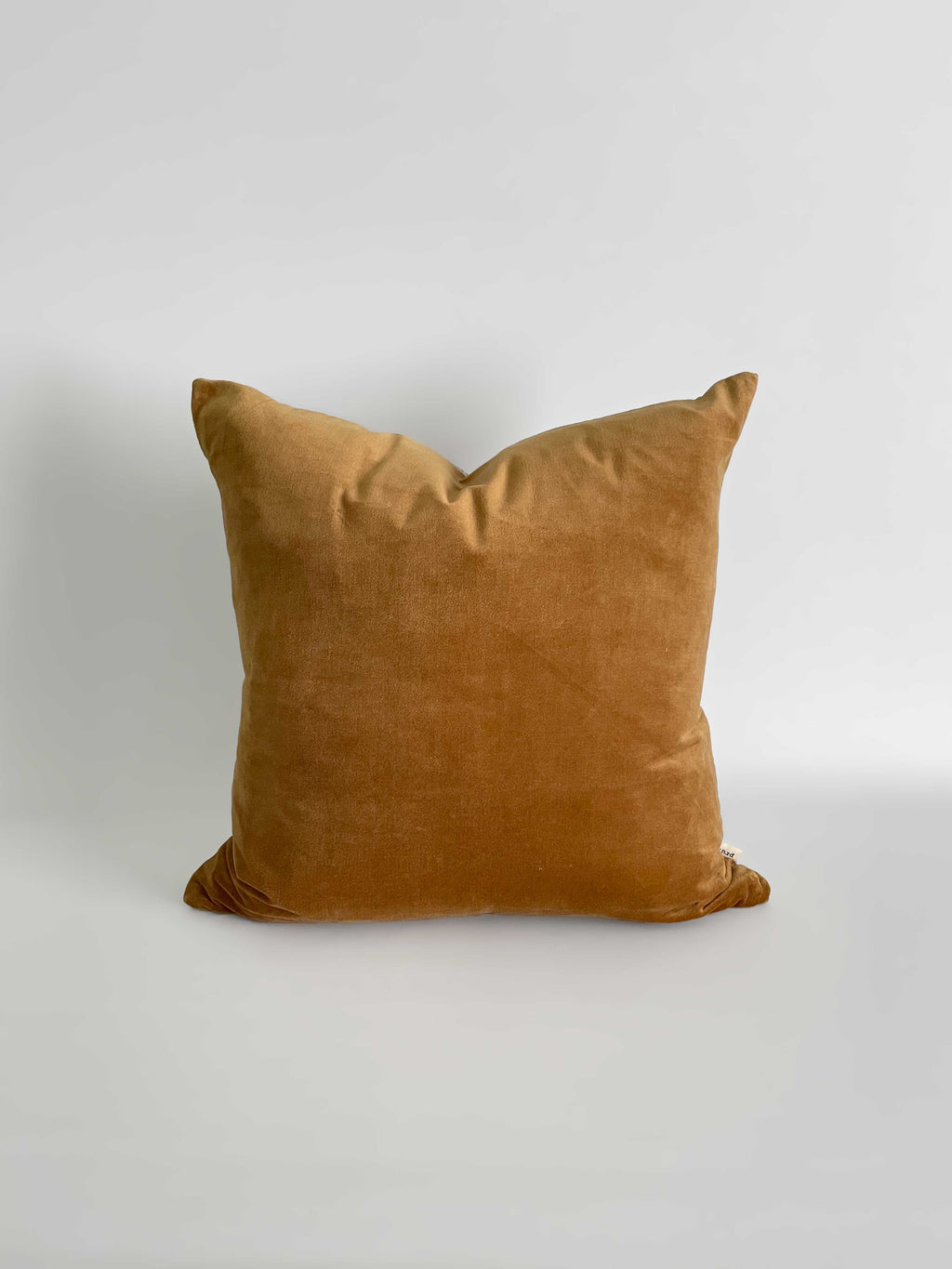 A square cushion in a warm caramel velvet fabric, against a neutral light background.