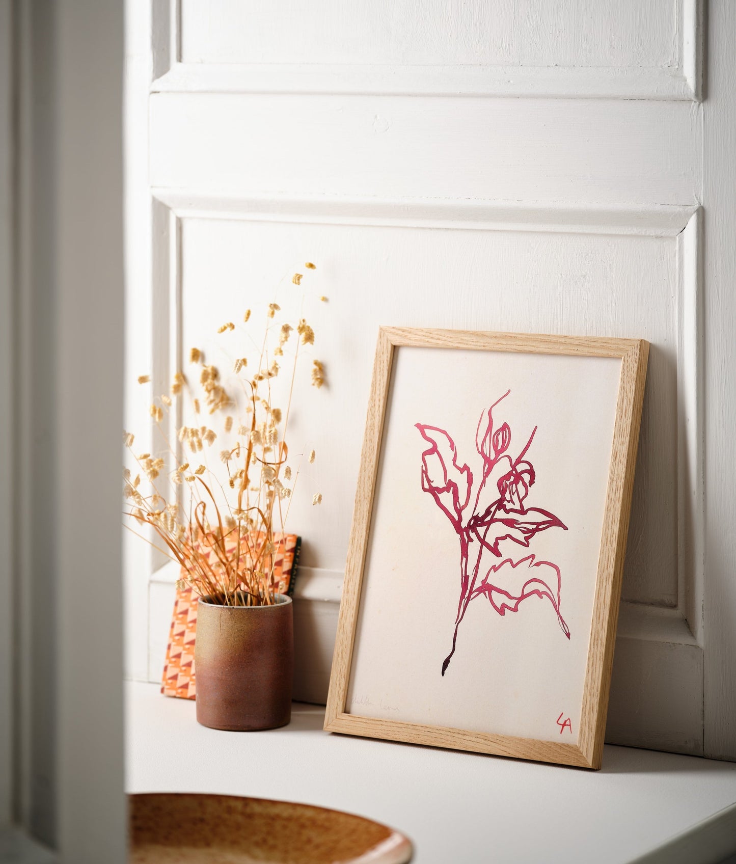 Art print of Dahlia leaves in red ink on vintage paper in a natural oak frame, sitting on a white painted windowsill alongside a stoneware vessel with dried flowers