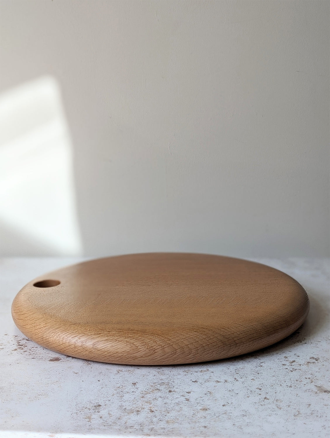 Large round English Oak serving board with hole for hanging, on an off-white, textured surface with a beige wall behind - side view.