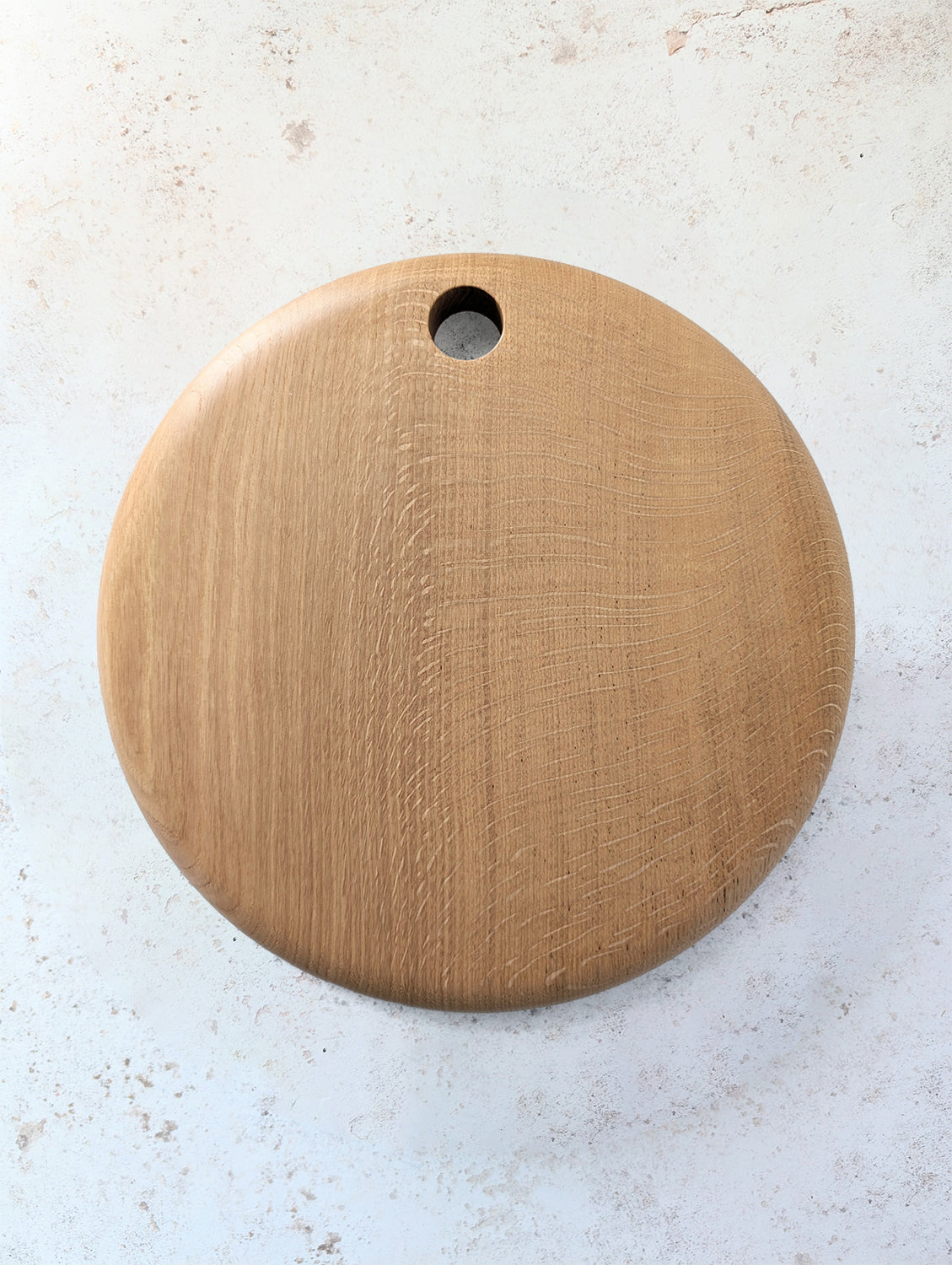 Large round English Oak serving board with hole for hanging, on an off-white, textured surface - top view.