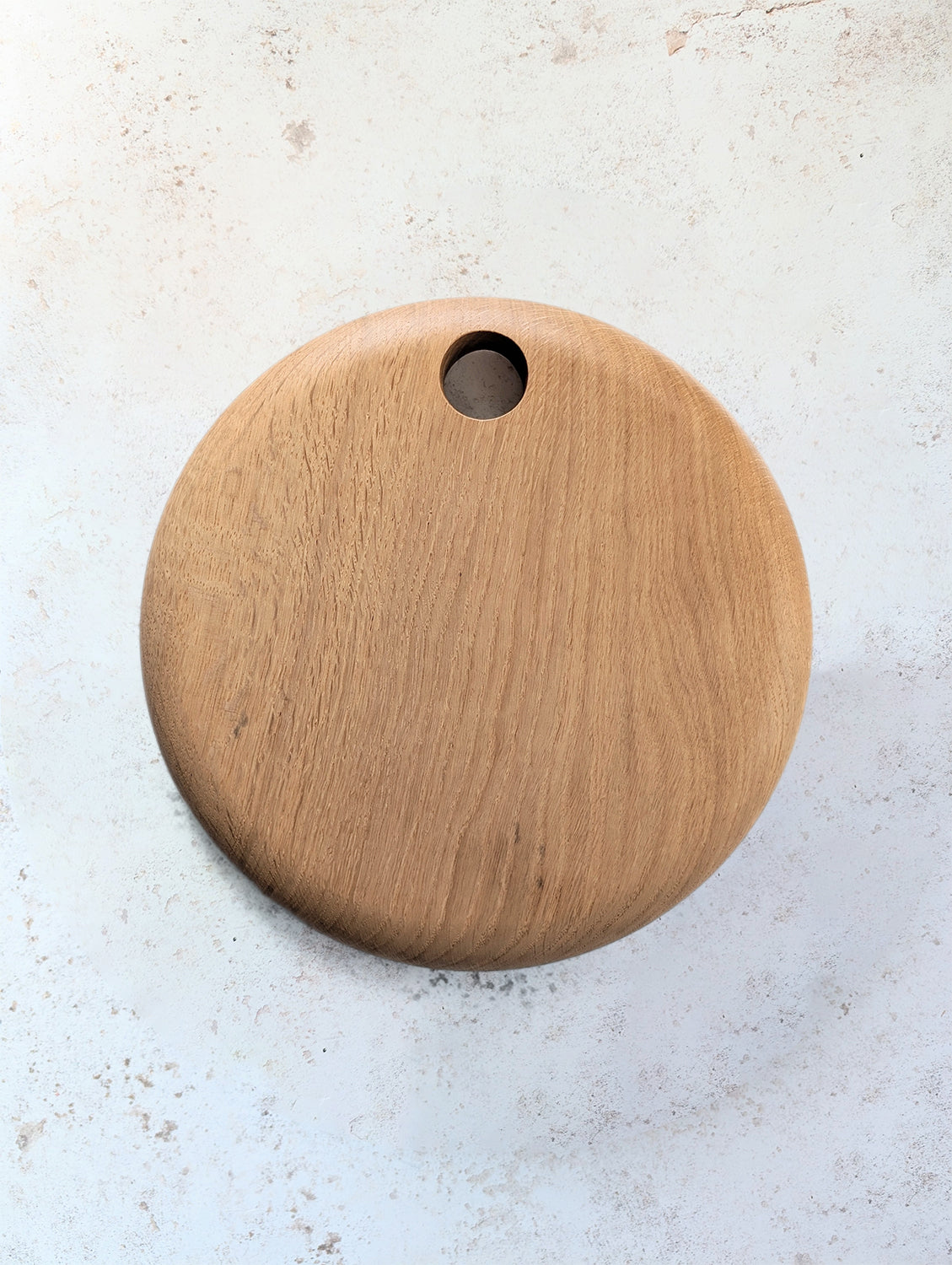 Medium round English Oak serving board with hole for hanging, on an off-white, textured surface - top view.