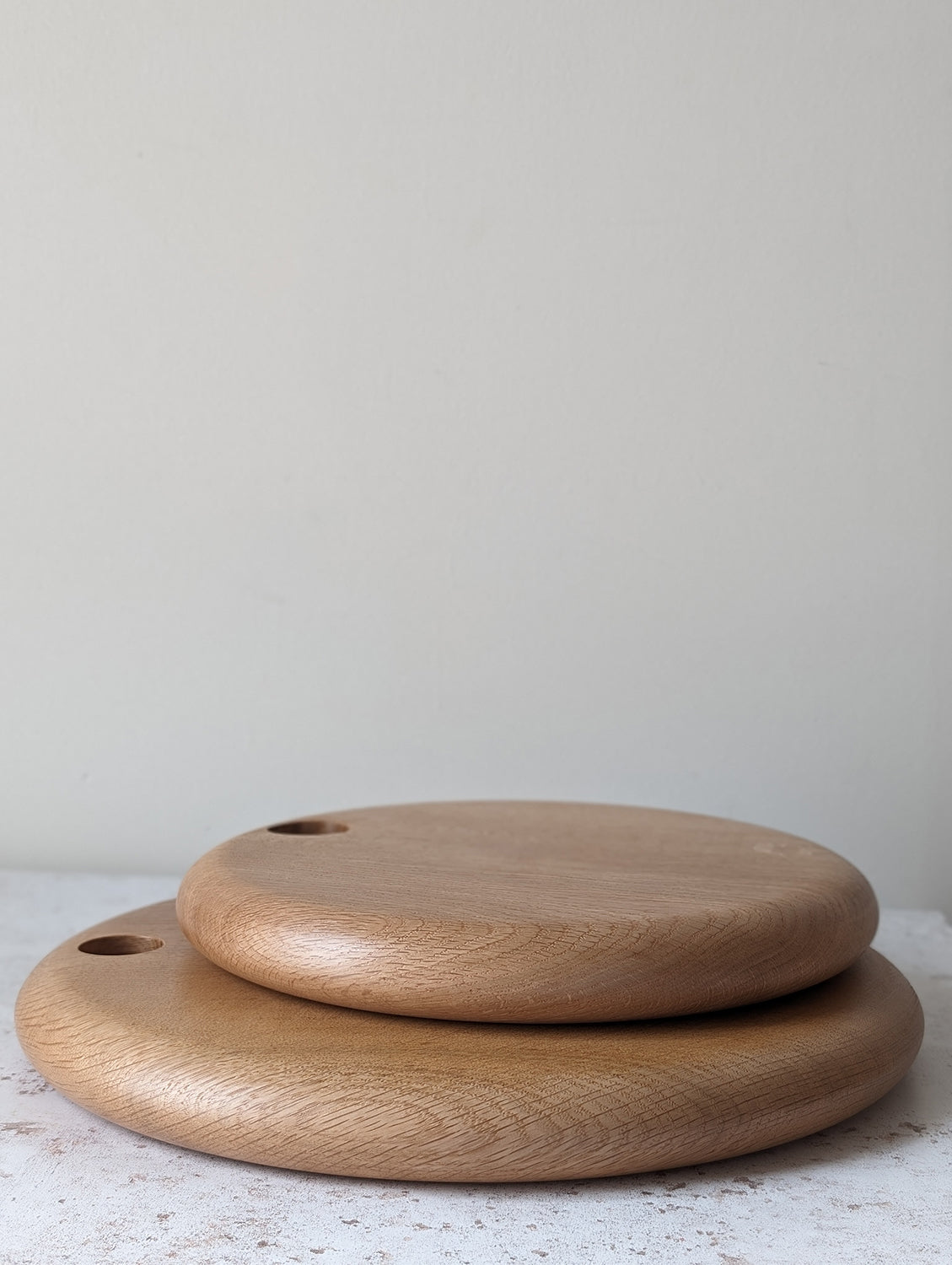 Pair of round English Oak serving boards with holes for hanging, stacked on an off-white, textured surface with a beige wall behind - side view.