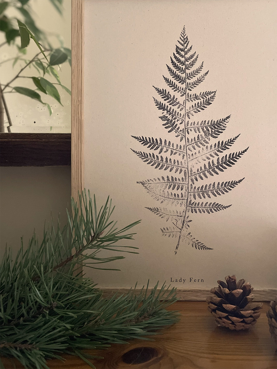 Framed print of a fern on a wall with greenery and pine cones in the foreground.