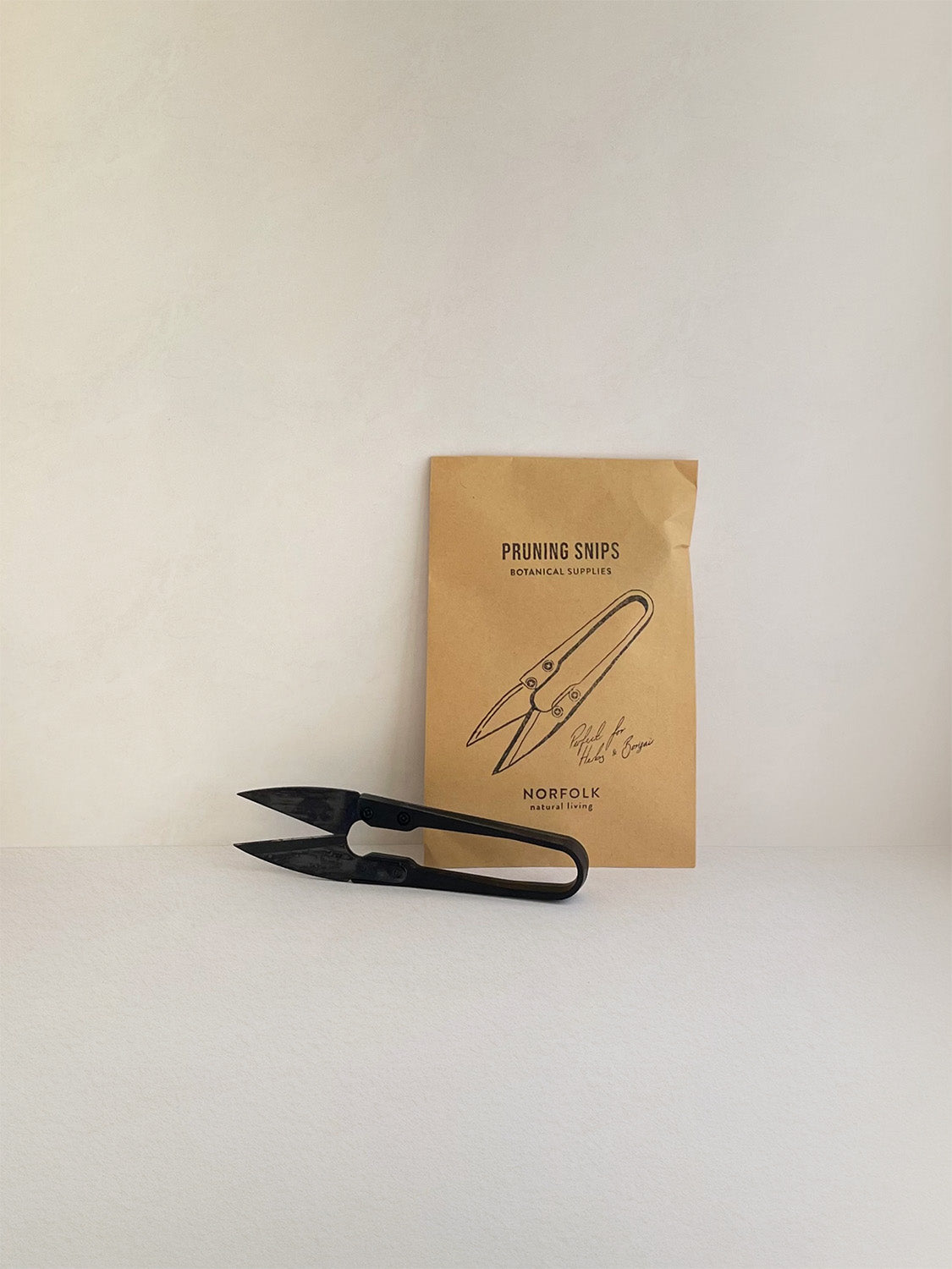A pair of black metal pruning snips resting in front of a small Kraft paper pouch with black text and illustration, on a neutral background.