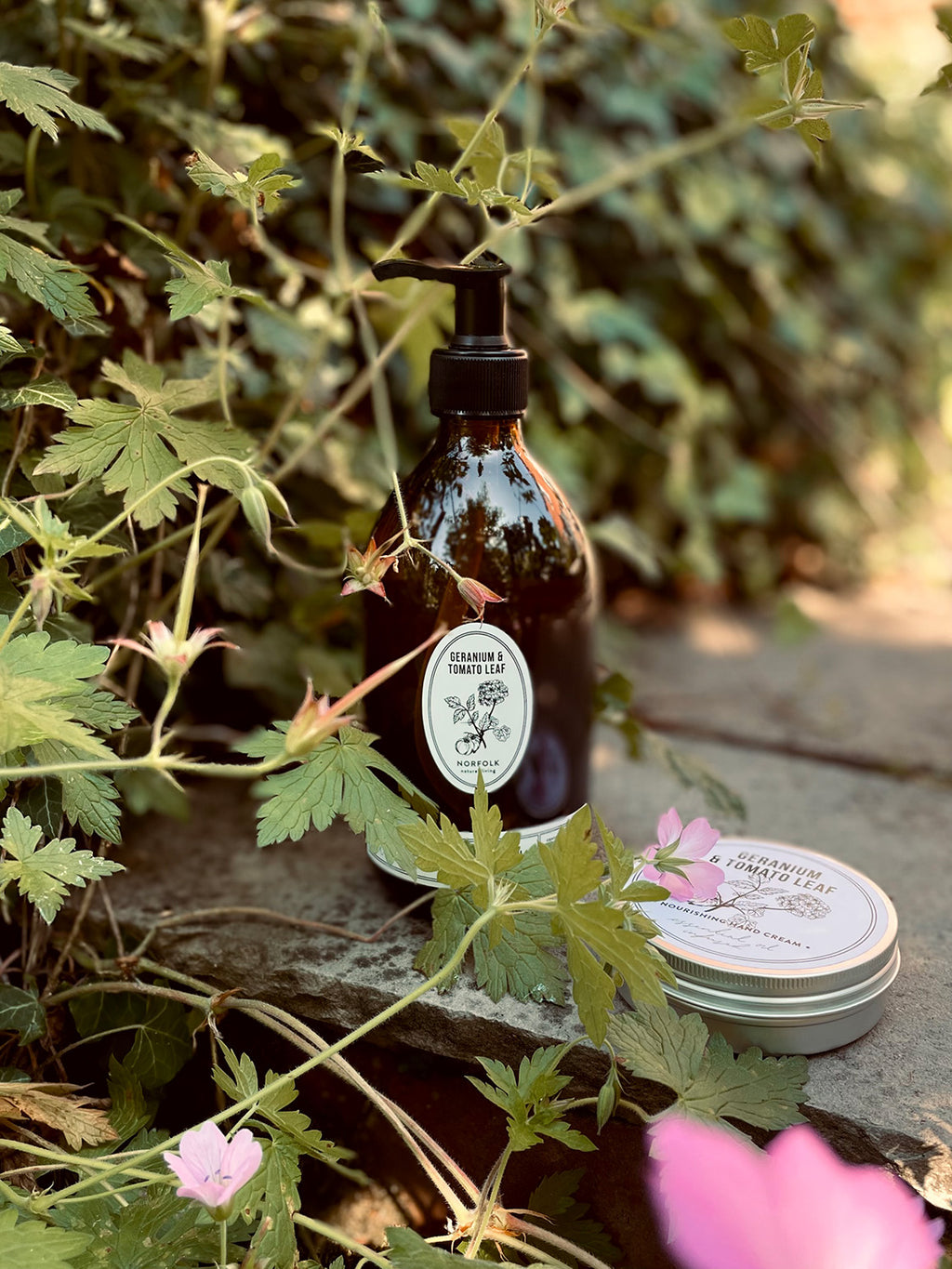 Geranium & Tomato Leaf hand wash bottle and hand cream tin with labels on a stone surface surrounded by greenery and flowers.