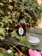 Geranium & Tomato Leaf hand wash bottle and hand cream tin with labels on a stone surface surrounded by greenery and flowers.