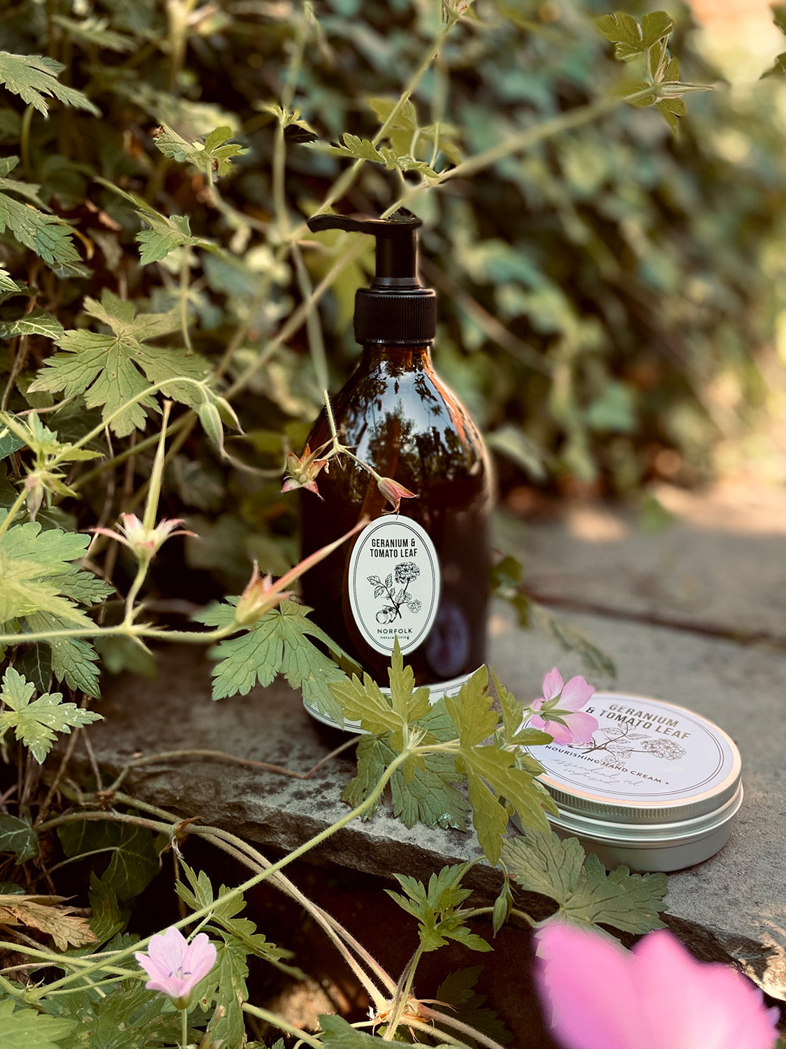 Geranium & Tomato Leaf hand wash bottle and hand cream tin with labels on a stone surface surrounded by greenery and flowers.