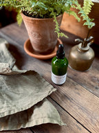 Indoor plant food solution in a dark green glass dropper bottle sitting on a rustic wooden surface, alongside a potted fern, vintage brass mister bottle and a folded linen cloth.