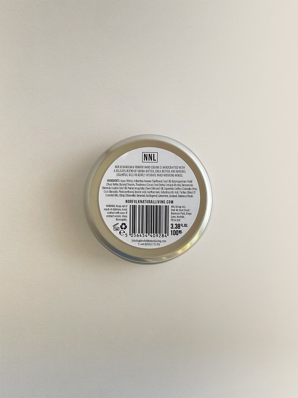 The back of a round metal tin of Geranium & Tomato Leaf nourishing hand cream, with a white label with black text, on a neutral background.